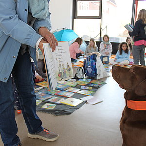 Welttag des Buches. Grundschüler veranstalten Bücherflohmarkt in der neuen Aula. Schulhund Carlos liest ein Buch.